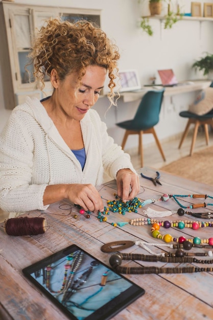 Une femme heureuse travaillant avec des perles et des cordons pour fabriquer des colliers et des accessoires apprenant à regarder un didacticiel en ligne sur une tablette avec connexion Internet Activité de loisir à l'intérieur Nouveau concept de travail dame