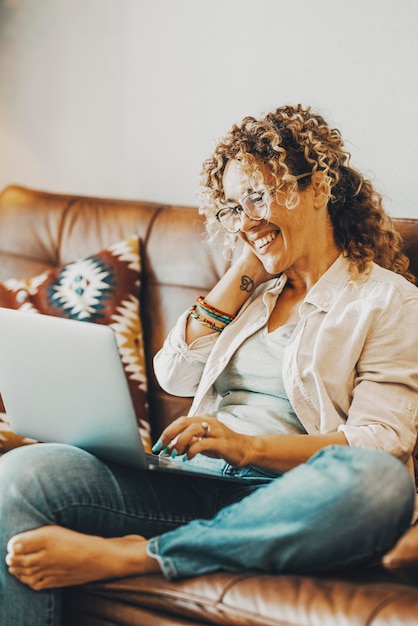 Femme heureuse utilisant un ordinateur portable à la maison pour appeler des amis ou des parents par vidéo assis sur le canapé et souriant Les femmes s'amusent avec le contenu de l'ordinateur et des médias sociaux La dame moderne aime écrire des pages Web