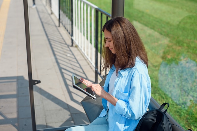 Femme heureuse utilise une tablette ou un ebook sur une station de tramway en attendant les transports publics.