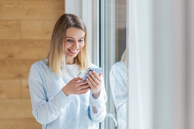 une femme heureuse a vu la bonne nouvelle sur un smartphone debout près de la fenêtre au bureau ou à la maison à l'intérieur