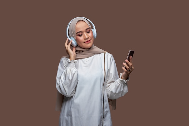 Femme en hijab portant des écouteurs sans fil tenant un smartphone tout en fermant les yeux