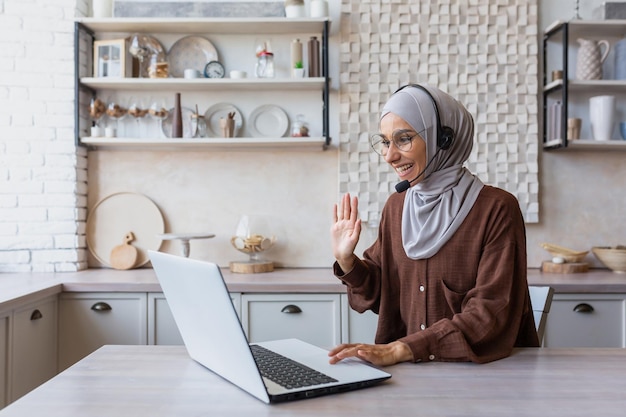Femme en hijab travaillant à distance depuis la maison assise dans la cuisine à l'aide d'un ordinateur portable et d'un casque pour un appel vidéo