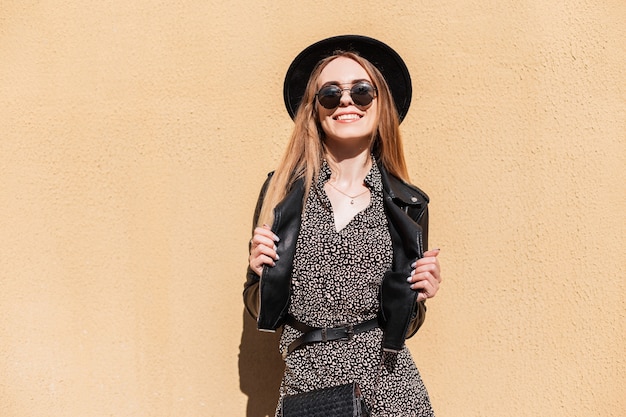 Femme hipster mode beauté avec un sourire parfait blanc dans des vêtements élégants au look d'automne avec des lunettes de soleil regardant la caméra près d'un mur par beau temps