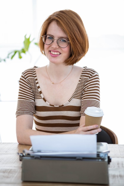 femme hipster souriante tenant une tasse de café, assis devant une machine à écrire