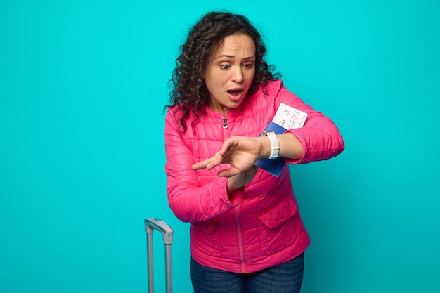 Photo une femme hispanique abasourdie en veste rose vif regarde sa montre avec surprise et peur, étant en retard pour son vol, tenant un passeport avec billet d'avion, debout à côté d'une valise sur fond bleu