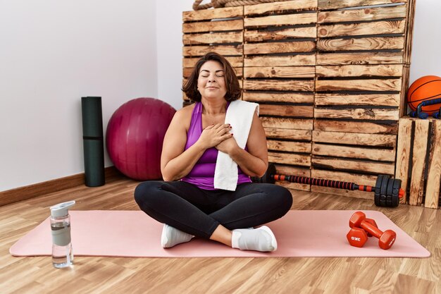 Femme hispanique d'âge moyen assise sur un tapis d'entraînement à la salle de gym souriante avec les mains sur la poitrine avec les yeux fermés et un geste reconnaissant sur le visage. notion de santé.