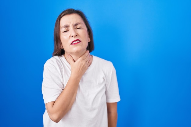 Femme hispanique d'âge moyen debout sur fond bleu touchant le cou douloureux mal de gorge pour la grippe et l'infection