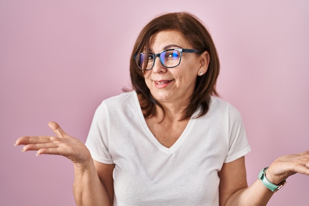 Une femme hispanique d'âge moyen debout sur un fond rose expression désemparée et confuse avec les bras et les mains a soulevé le concept de doute