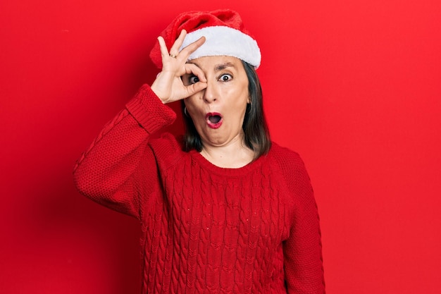 Femme hispanique d'âge moyen portant un chapeau de noël faisant un geste ok choqué par l'œil du visage surpris regardant à travers les doigts expression incrédule