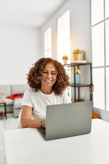 Femme hispanique d'âge moyen souriant confiant utilisant un ordinateur portable à la maison