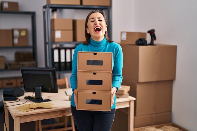 Femme hispanique d'âge moyen travaillant dans une petite entreprise de commerce électronique tenant des boîtes souriant et riant fort à haute voix parce qu'une drôle de blague folle