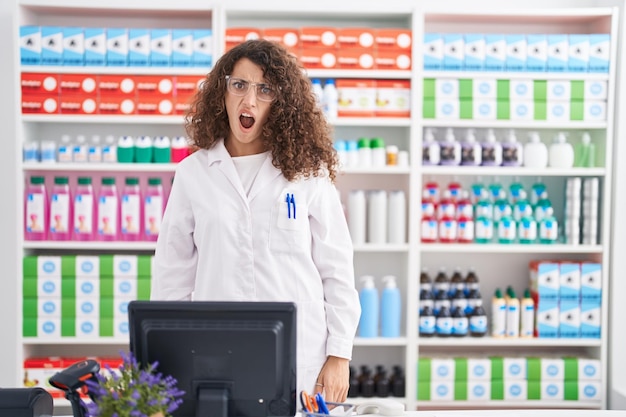 Femme hispanique aux cheveux bouclés travaillant à la pharmacie pharmacie en état de choc à la sceptique et sarcastique surpris avec la bouche ouverte