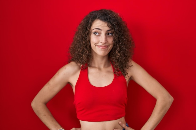 femme hispanique, à, cheveux bouclés, debout, sur, arrière-plan rouge, sourire, regarder côté, et, regarder loin, pensée