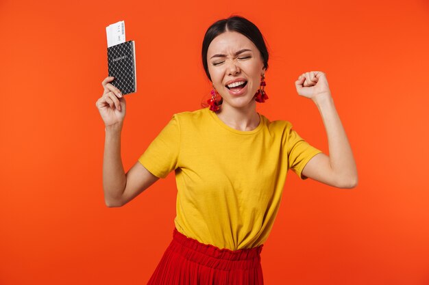 femme hispanique émotionnelle des années 20 vêtue d'une jupe souriante tout en tenant un passeport et des billets de voyage isolés sur un mur rouge