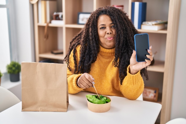 Femme hispanique de grande taille mangeant une salade à emporter tenant un smartphone souriant regardant sur le côté et regardant loin de penser.