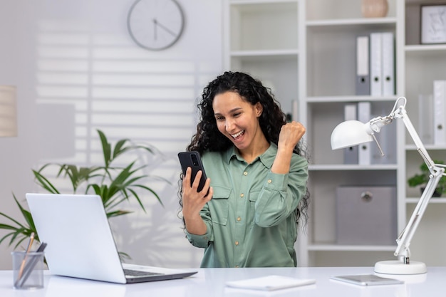 Une femme hispanique heureuse assise au bureau à la table se rassemblant en regardant l'écran du mobile