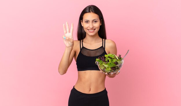 Femme hispanique de remise en forme souriante et amicale montrant le numéro trois et tenant une salade