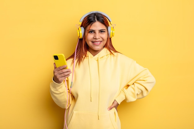 Femme hispanique souriant joyeusement avec une main sur la hanche et confiante. casque et concept de smartphone