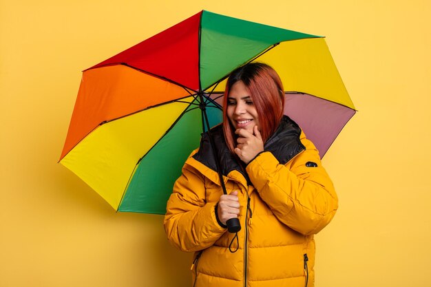 Femme hispanique souriante avec une expression heureuse et confiante avec la main sur le menton. concept de pluie et de parapluie