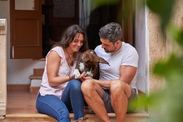 Femme et homme aimants en vêtements décontractés assis dans les escaliers à l'extérieur de la maison et étreignant un adorable chien ensemble