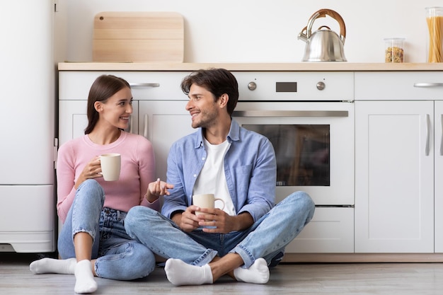 Une femme et un homme caucasiens souriants et heureux du millénaire s'assoient sur le sol avec des tasses de boisson chaude parlant dans une cuisine moderne