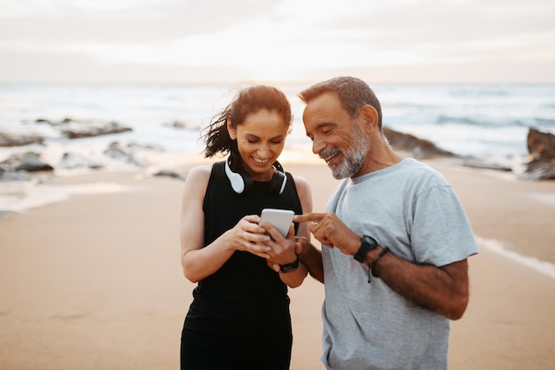 Une femme et un homme européens matures souriants en vêtements de sport se reposent de l'entraînement avec un smartphone