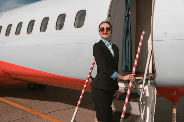 Femme hôtesse de l'air à lunettes de soleil debout sur les escaliers de l'avion à l'aéroport et regardant au loin