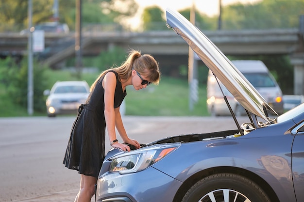 Femme impuissante debout près de sa voiture avec capot ouvert inspectant un moteur cassé Jeune conductrice ayant des problèmes avec le véhicule