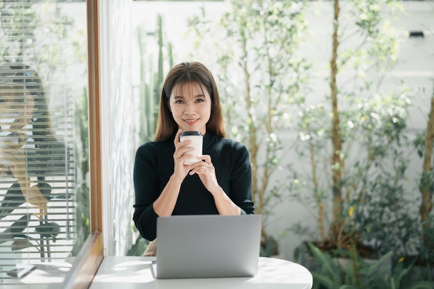 Femme indépendante asiatique souriante tenant une tasse de café chaud et travaillant sur un ordinateur portable sur une table en bois au café Femme entrepreneur travaillant pour son entreprise au café Travail d'entreprise de n'importe où