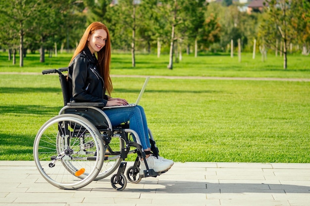 Femme indépendante au gingembre rousse assise dans un fauteuil roulant et travaillant par ordinateur portable dans un parc d'été.