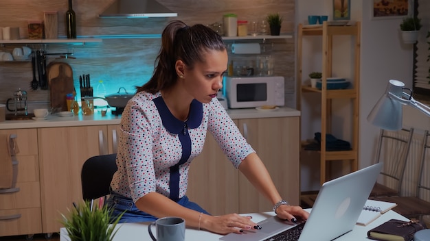 Femme indépendante concentrée travaillant des heures supplémentaires pour terminer un projet de cuisine à domicile. Employé concentré occupé utilisant un réseau de technologie moderne sans fil faisant des heures supplémentaires pour la lecture d'emplois, la saisie, la recherche