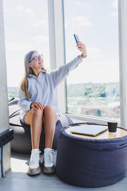 Femme indépendante dans des verres classiques parlant au téléphone et souriant assis dans un espace de coworking moderne femme millénaire insouciante dans des verres profitant du temps libre pour la communication