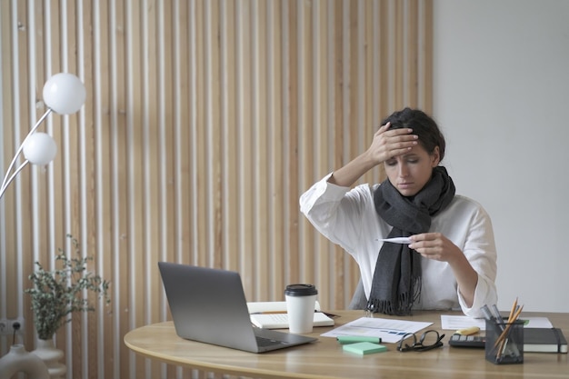 Femme indépendante fatiguée assise au bureau au bureau à domicile regardant maladivement un thermomètre numérique