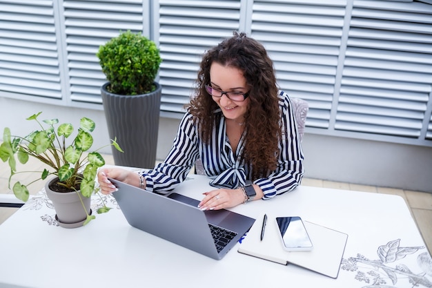Femme indépendante pensive dans des vêtements décontractés à la mode travaillant avec un ordinateur portable et prenant des notes, souriant et regardant la terrasse extérieure du café