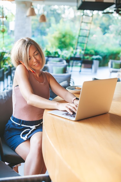 Femme indépendante souriante parlant par téléphone portable, assise dans le café extérieur ensoleillé, travaillant sur un ordinateur portable. Travail de bureau confortable, travail à distance et concept de travail à domicile.