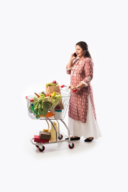 Femme indienne d'âge moyen avec panier ou chariot plein d'épicerie, de légumes et de fruits. Photo pleine longueur isolée sur mur blanc