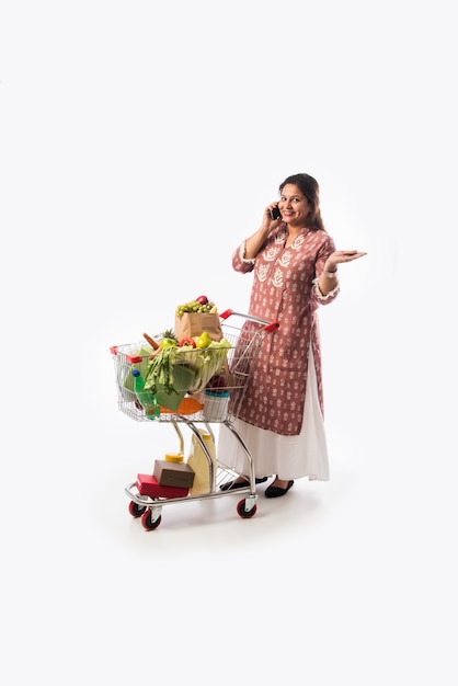 Femme indienne d'âge moyen avec panier ou chariot plein d'épicerie, de légumes et de fruits. Photo pleine longueur isolée sur mur blanc
