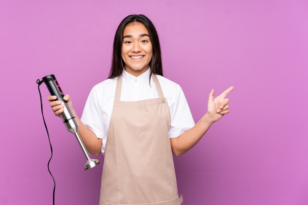 Femme indienne à l'aide d'un mixeur plongeant isolé sur fond violet doigt pointé sur le côté