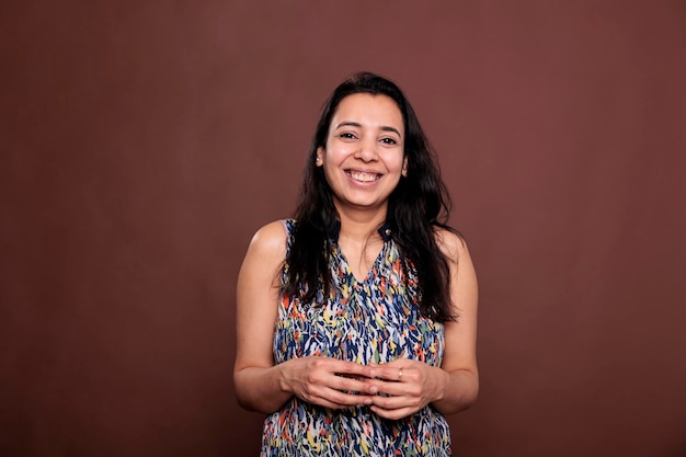 Femme indienne souriante entrelacant le portrait des doigts, riant, regardant la caméra. Dame positive, jeune personne debout avec une expression faciale joyeuse, vue de face en studio