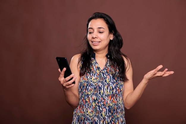 Une femme indienne souriante envoie des messages sur les réseaux sociaux, regarde des vidéos en ligne sur un smartphone, lit sur Internet. Dame joyeuse tenant un téléphone portable, textos, vue de face studio moyen plan