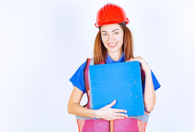 Femme ingénieur en casque rouge tenant un dossier de projet bleu.