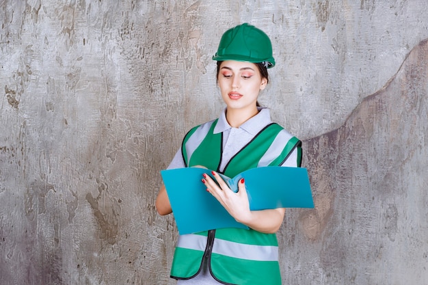 Femme ingénieur en casque vert tenant un dossier bleu.