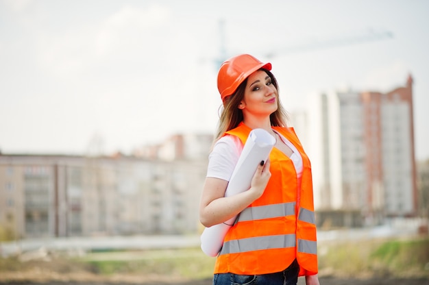 Femme ingénieur constructeur en gilet uniforme et casque de protection orange détiennent des documents commerciaux contre les nouveaux bâtiments avec grue.