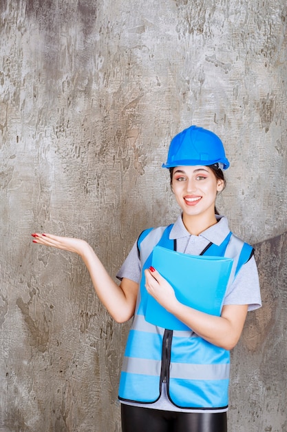 Femme ingénieur en uniforme bleu et casque tenant un dossier de rapport bleu et pointant vers quelqu'un autour