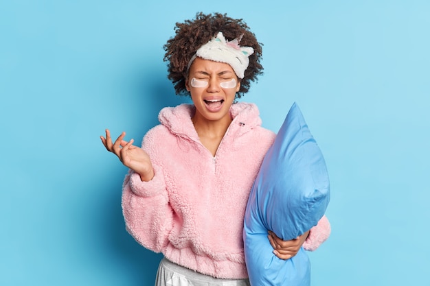 Une femme insatisfaite pleure de désespoir vêtue d'un masque de sommeil pyjama applique des patchs d'hydrogel sous les yeux tient un oreiller doux va au lit isolé sur un mur bleu