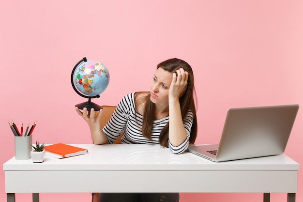 Une femme insatisfaite se penche sur la main tenant un globe ayant des problèmes avec la planification des vacances alors qu'elle est assise, travaille au bureau avec un ordinateur portable isolé sur fond rose pastel. Carrière commerciale de réussite. Espace de copie.