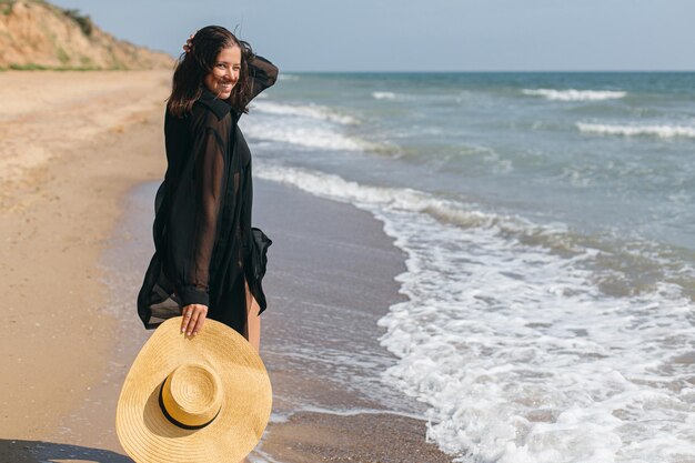 Femme insouciante avec un chapeau marchant sur une plage de sable au bord de la mer et des vacances d'été relaxantes
