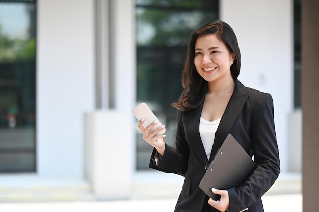 Femme intelligente utilisant un téléphone intelligent pour discuter de la communication d'entreprise tout en marchant dans le quartier des bureaux modernes