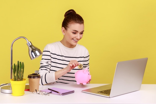 Femme investissant dans des bitcoins mettant une pièce de monnaie crypto dorée dans une tirelire et regardant l'écran d'un ordinateur portable