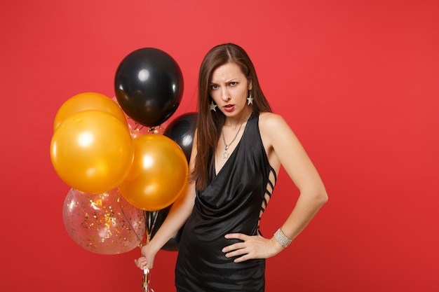 Femme irritée en colère en robe noire tenant des ballons à air debout dans une pose menaçante tout en célébrant isolé sur fond rouge. Saint Valentin Happy New Year, concept de fête de vacances de maquette d'anniversaire.